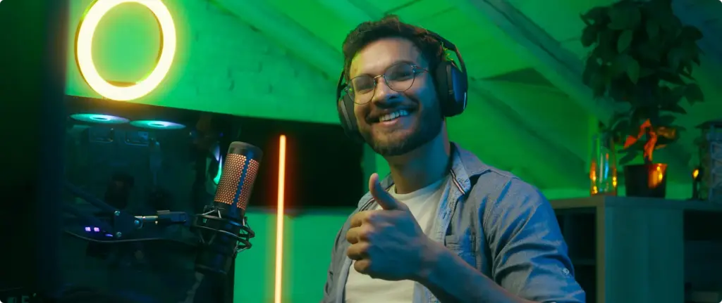 Een man met een koptelefoon op glimlacht en steekt zijn duim omhoog terwijl hij achter een computer met gamingapparatuur zit. De kamer wordt verlicht door groene en oranje neonlichten, wat voor een levendige sfeer zorgt
